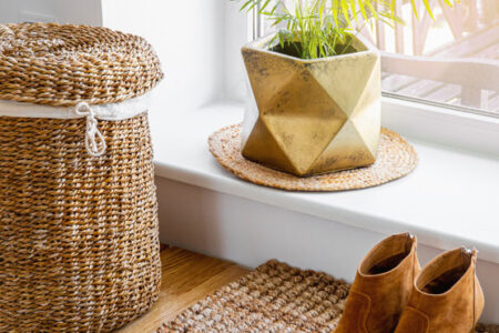 Hardwood floor with jute doormat, shoes and flower pot and seagrass laundry basket by window. Natural material objects in home concept. Home interior.
